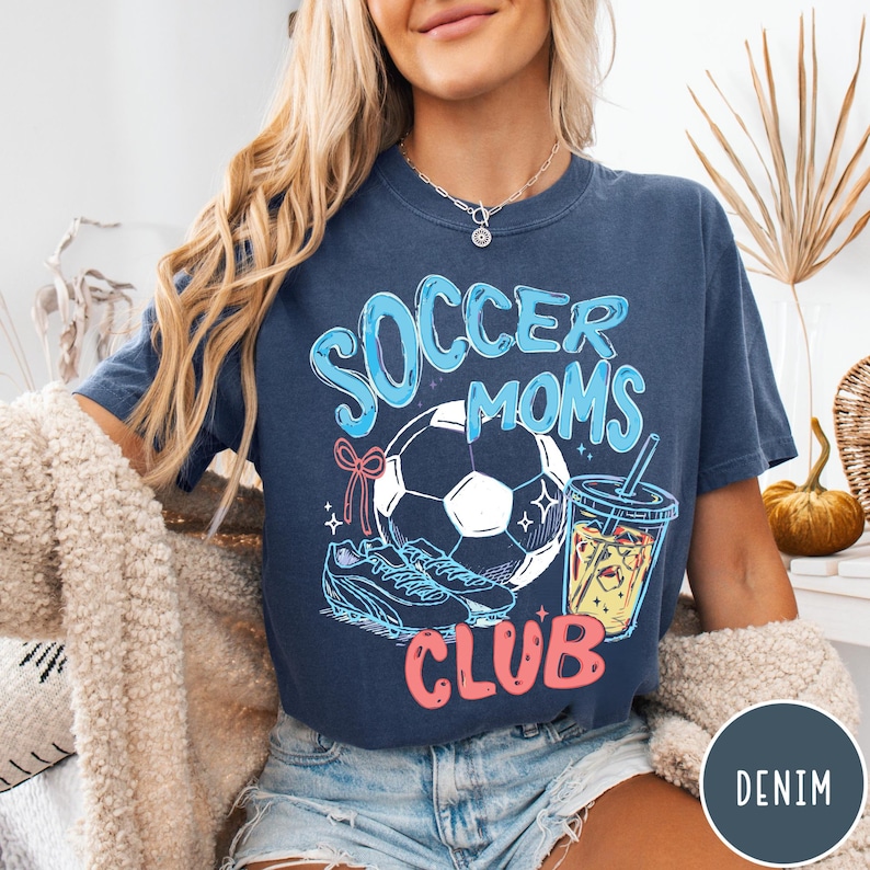 May include: A navy blue t-shirt with the words "Soccer Moms Club" in a playful font. The graphic includes a soccer ball, cleats, and a drink. The shirt is a comfortable, casual style.