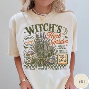 May include: An ivory t-shirt featuring a graphic print with the text "WITCH'S Herb Garden" and "FRESHLY GATHERED". The design includes illustrations of a bundle of herbs, mushrooms, a potion bottle, a crescent moon, and the words "SAGE POTIONS MOONLIGHT".