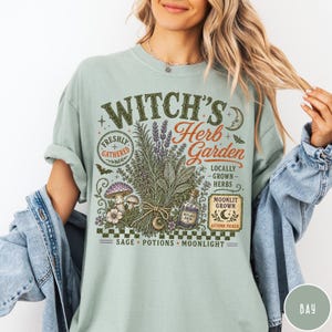 May include: A sage green t-shirt featuring a graphic print with the text "Witch's Herb Garden" and "Locally Grown Herbs." The design includes illustrations of a bundle of herbs, mushrooms, a potion bottle, a crescent moon, and a checkerboard pattern.