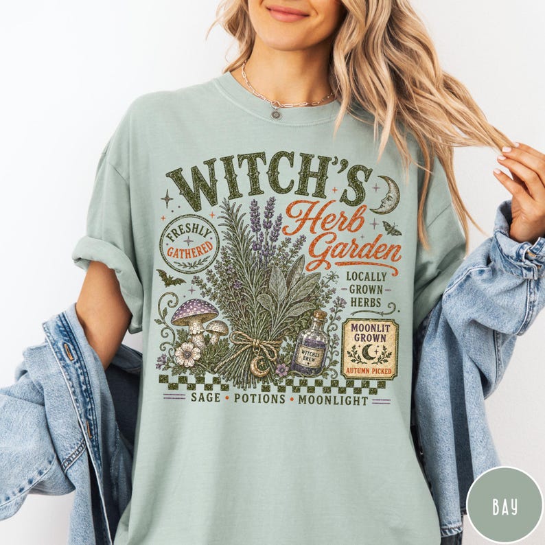 May include: A sage green t-shirt featuring a graphic print with the text "Witch's Herb Garden" and "Locally Grown Herbs." The design includes illustrations of a bundle of herbs, mushrooms, a potion bottle, a crescent moon, and a checkerboard pattern.