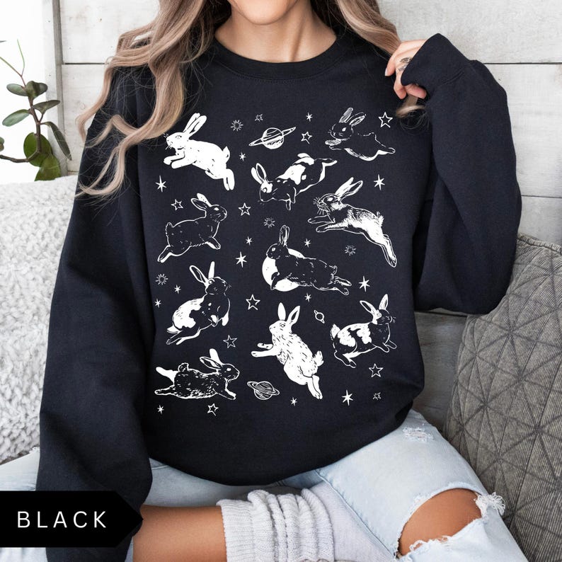 May include: Black sweatshirt featuring a white print of rabbits, planets, and stars. The word "BLACK" is printed at the bottom left. The sweatshirt has a crew neck design.
