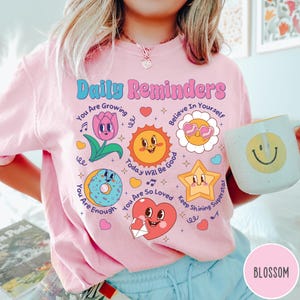 May include: Pink sweatshirt with the text "Daily Reminders" and various cartoon graphics, including a tulip, sun, flower, donut, star, and heart. The sweatshirt also features the words "You Are Growing", "Believe In Yourself", "You Are Enough", "Today Will Be Good", "You Are So Loved", and "Keep Shining Superstar".