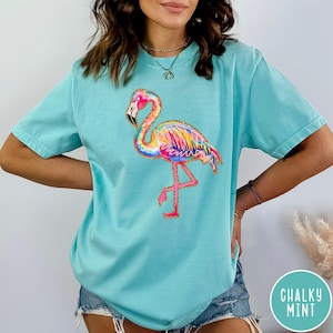May include: A light blue t-shirt featuring a colorful flamingo design. The flamingo has pink, yellow, blue, and orange feathers with gold glitter accents. The shirt is a comfortable, casual style.