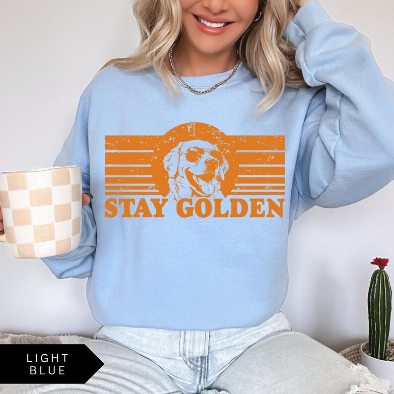 Puede incluir: Una sudadera de color azul claro con un gr&aacute;fico retro de un golden retriever con gafas de sol. El gr&aacute;fico dice "Stay Golden" en texto naranja.