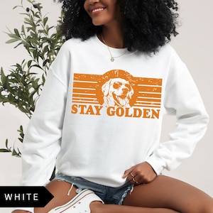 Puede incluir: Sudadera blanca con un gr&aacute;fico naranja de un golden retriever con gafas de sol y el texto "Stay Golden".