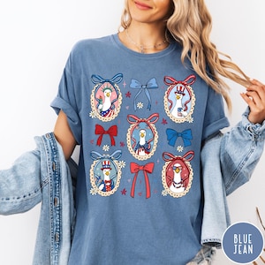 May include: A blue t-shirt featuring a patriotic design with cartoon geese wearing hats and holding items like the Statue of Liberty torch. The shirt has a round neck and short sleeves. The design includes red, white, and blue bows and floral accents.