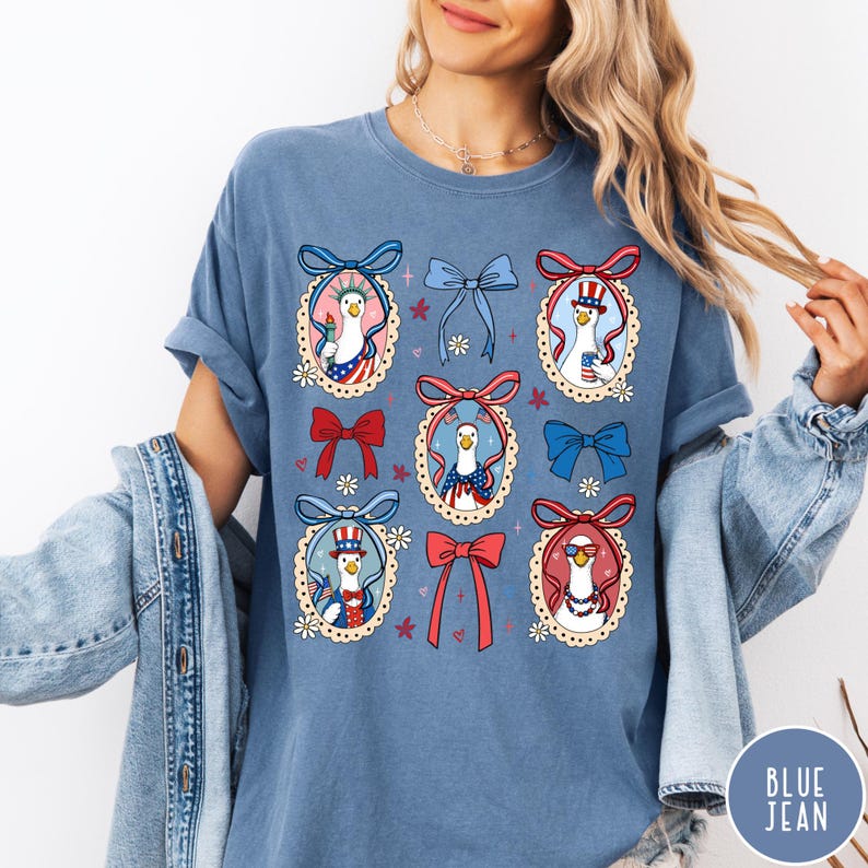 May include: A blue t-shirt featuring a patriotic design with cartoon geese wearing hats and holding items like the Statue of Liberty torch. The shirt has a round neck and short sleeves. The design includes red, white, and blue bows and floral accents.
