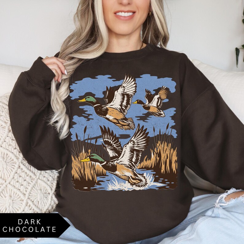 May include: Dark chocolate-colored sweatshirt featuring a graphic of three ducks in flight over a body of water with reeds and a blue sky background. The text "DARK CHOCOLATE" is at the bottom.