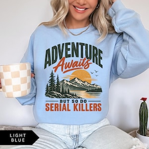 May include: A light blue crewneck sweatshirt featuring a graphic print with the text ADVENTURE Awaits BUT SO DO SERIAL KILLERS. The design includes a dark green forest, a large orange sun, and a mountain range reflected in a body of water.