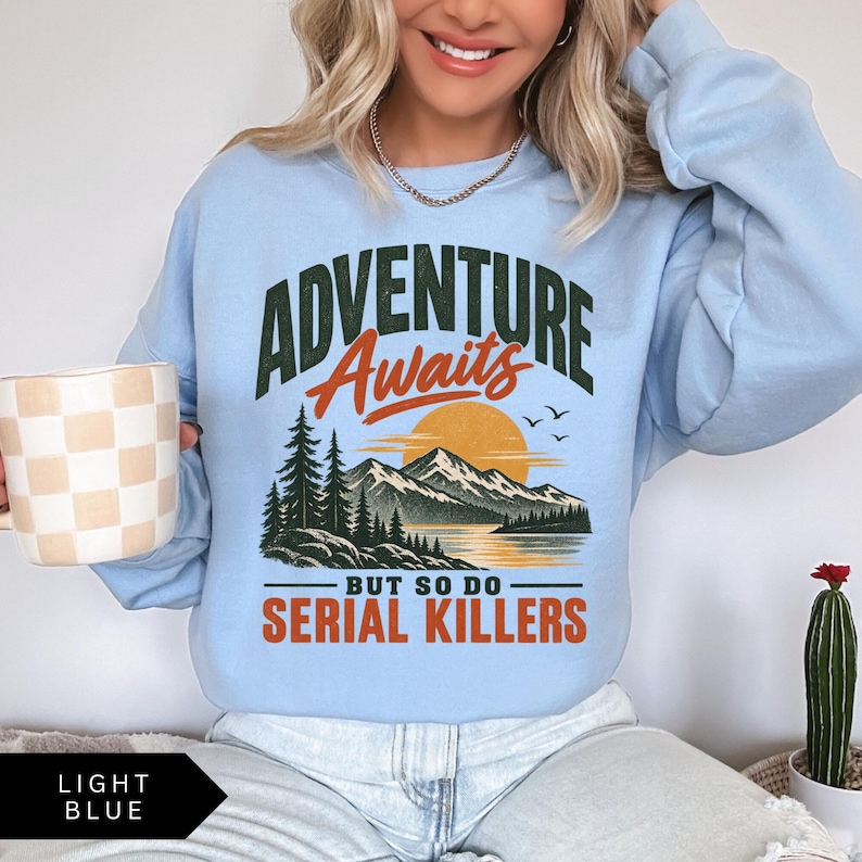 May include: A light blue crewneck sweatshirt featuring a graphic print with the text ADVENTURE Awaits BUT SO DO SERIAL KILLERS. The design includes a dark green forest, a large orange sun, and a mountain range reflected in a body of water.