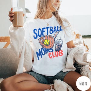May include: White t-shirt with a graphic design featuring a softball glove, ball, and iced drink. The text reads "SOFTBALL MOMS CLUB" in blue and red. The shirt is a casual style.