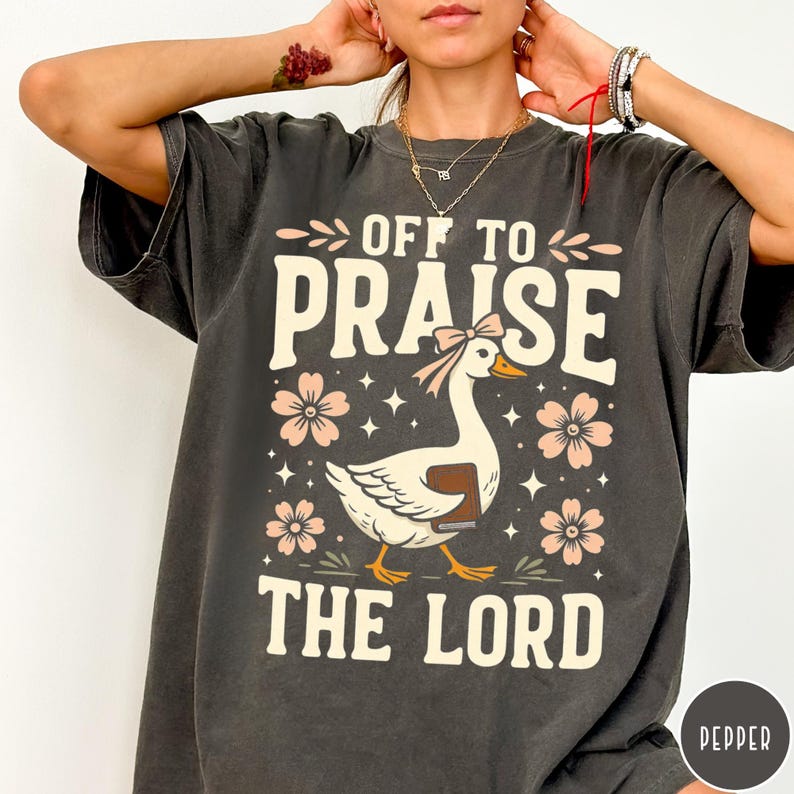 Puede incluir: Camiseta gris oscuro con el texto &laquo;OFF TO PRAISE THE LORD&raquo;. La camiseta presenta un ganso blanco con un libro, flores rosas y estrellas. El ganso tiene un lazo rosa en la cabeza.
