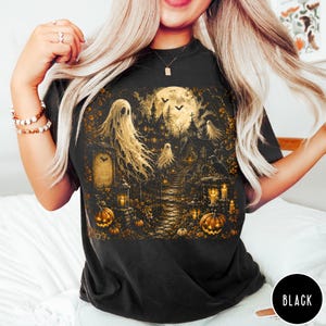 Puede incluir: Una camiseta negra con un estampado gr&aacute;fico detallado de una escena embrujada con fantasmas, luna llena, una mansi&oacute;n oscura, calabazas y linternas. La ilustraci&oacute;n utiliza tonos dorados y marrones c&aacute;lidos para representar el cementerio.