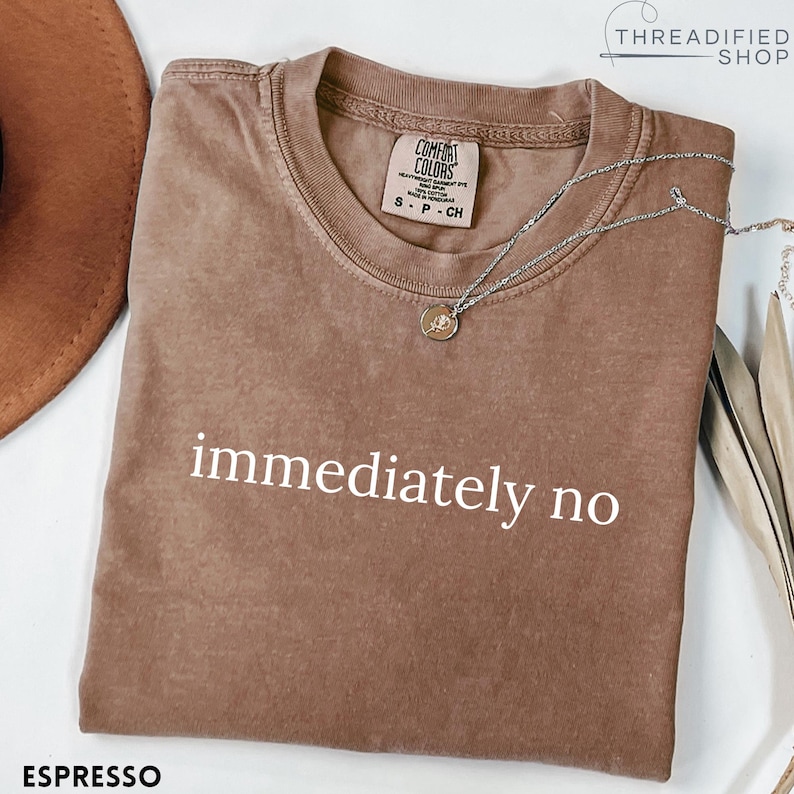 May include: Espresso-colored t-shirt with the phrase "immediately no" in white. The shirt has a crew neck and short sleeves. A silver necklace with a small pendant rests on the shirt. The brand tag is visible.