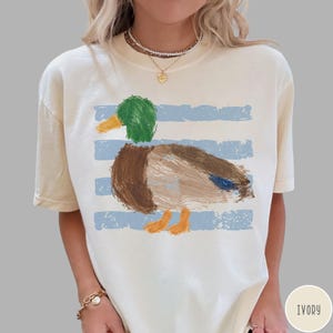 May include: An ivory short-sleeve t-shirt featuring a painterly graphic of a mallard duck with a green head, brown body, and orange feet. The duck is positioned over horizontal light blue distressed stripes. The shirt has a crew neckline.