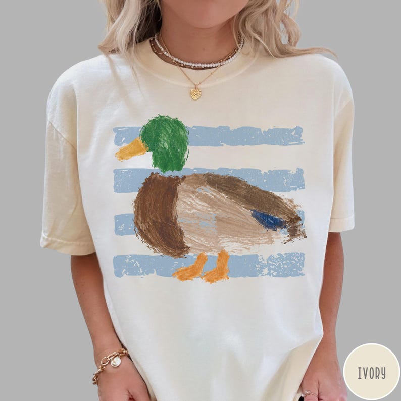May include: An ivory short-sleeve t-shirt featuring a painterly graphic of a mallard duck with a green head, brown body, and orange feet. The duck is positioned over horizontal light blue distressed stripes. The shirt has a crew neckline.