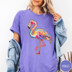 May include: Violet-colored t-shirt featuring a colorful flamingo design. The flamingo has a pink head and legs, a black beak, and a body with rainbow-colored feathers outlined in gold glitter. The shirt is a comfortable, casual style.