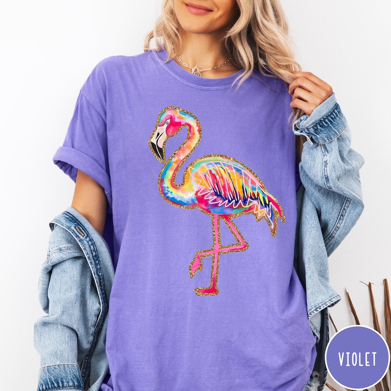 May include: Violet-colored t-shirt featuring a colorful flamingo design. The flamingo has a pink head and legs, a black beak, and a body with rainbow-colored feathers outlined in gold glitter. The shirt is a comfortable, casual style.