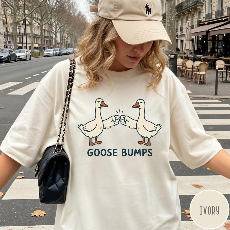 Puede incluir: Una camiseta de manga corta color marfil con un gr&aacute;fico de dos gansos blancos d&aacute;ndose un choque de pu&ntilde;os. Debajo de los gansos, el texto "GOOSE BUMPS" aparece en letras may&uacute;sculas de color azul oscuro. Se combina con un bolso negro acolchado.