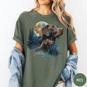 May include: Moss green t-shirt featuring a watercolor illustration of two brown dogs with a full moon and forest background. The shirt has short sleeves and a relaxed fit. The word "MOSS" is printed on a green circle.