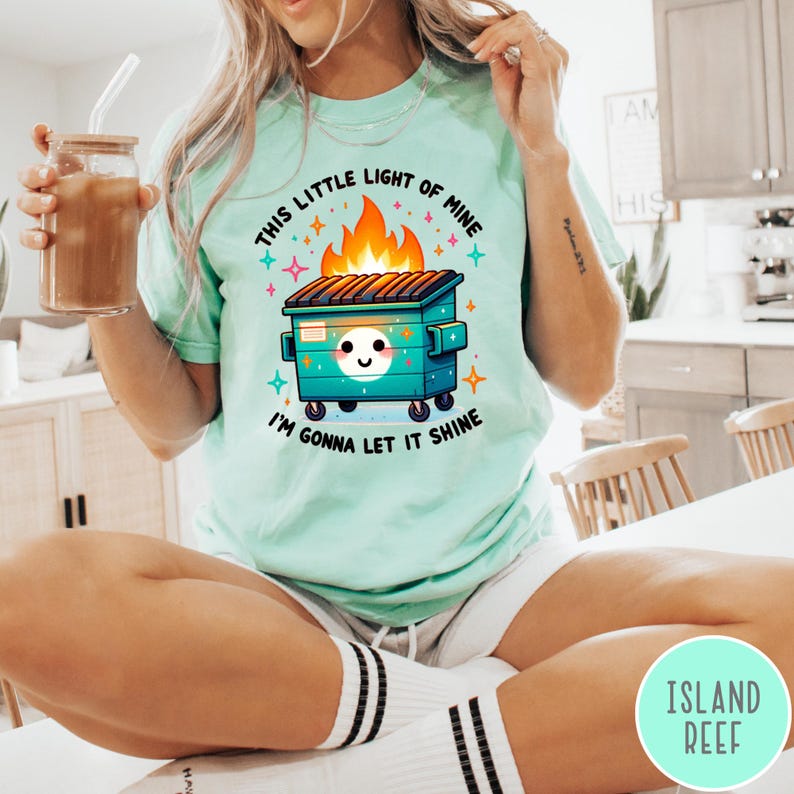 May include: A light green t-shirt with a graphic of a cartoon dumpster on fire with the text "This little light of mine I'm gonna let it shine".