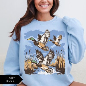 May include: Light blue sweatshirt featuring a graphic of three ducks in flight over water and reeds. The ducks have brown, white, and green details. The text "LIGHT BLUE" is in a black box at the bottom left.