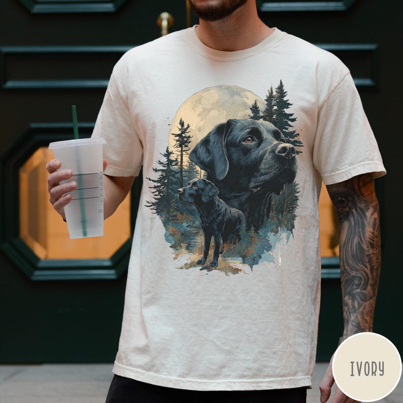May include: Ivory-colored t-shirt featuring a detailed illustration of a black Labrador Retriever with a full moon and forest background. The design includes two Labrador portraits, one large and one smaller, with a serene, naturalistic aesthetic.