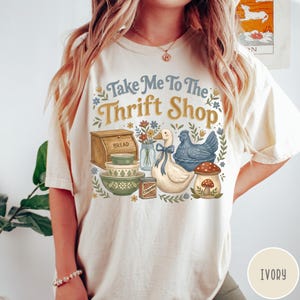 Op de afbeelding: Ivoor kleurig T-shirt met de tekst "Take Me To The Thrift Shop". De afbeelding toont een brooddoos, kommen, een eend met een blauw lint, een blauwe kip en een paddenstoel. Bloemen en bladeren omlijsten de illustraties.