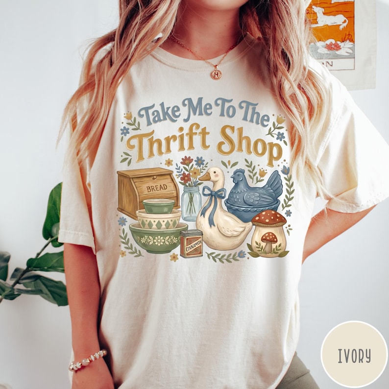 Op de afbeelding: Ivoor kleurig T-shirt met de tekst "Take Me To The Thrift Shop". De afbeelding toont een brooddoos, kommen, een eend met een blauw lint, een blauwe kip en een paddenstoel. Bloemen en bladeren omlijsten de illustraties.