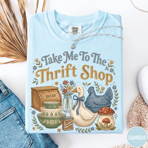 Op de afbeelding: Lichtblauw Comfort Colors T-shirt met de tekst "Take Me To The Thrift Shop" en illustraties van een brooddoos, kommen, een eend, een kip en een paddenstoel. Een zilveren ketting ligt over het shirt.