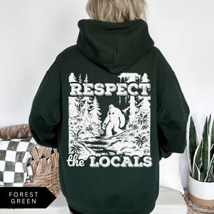 May include: A dark green hooded sweatshirt with a white graphic print of a Bigfoot in a forest setting. The text "Respect the Locals" is printed below the image.