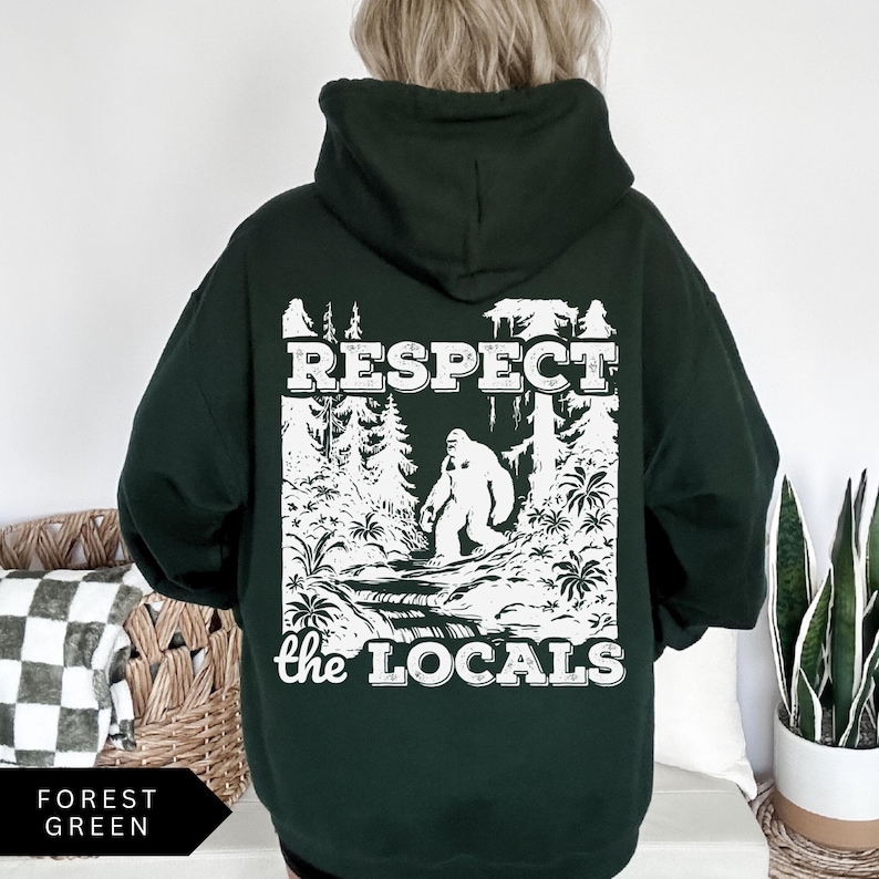 May include: A dark green hooded sweatshirt with a white graphic print of a Bigfoot in a forest setting. The text "Respect the Locals" is printed below the image.