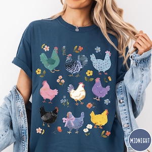 K&ouml;nnte beinhalten: Ein marineblaues T-Shirt mit einem bunten Muster aus H&uuml;hnern und Blumen. Die H&uuml;hner sind alle in verschiedenen Farben und haben unterschiedliche Muster. Die Blumen sind rosa, gelb und lila.