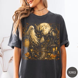 Puede incluir: Una camiseta gris carb&oacute;n con un estampado gr&aacute;fico detallado de una escena embrujada con fantasmas, luna llena, una mansi&oacute;n oscura, murci&eacute;lagos y calabazas naranjas brillantes, un camino de piedra, linternas y hojas de oto&ntilde;o.
