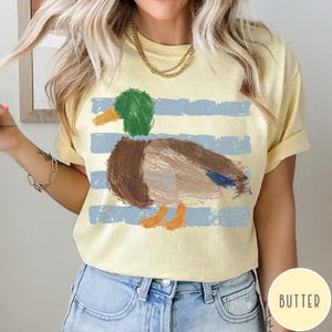 May include: A pale yellow short-sleeve t-shirt featuring a graphic of a mallard duck with a green head, brown body, and orange feet. The duck is positioned over horizontal, distressed light blue stripes. The shirt has a crew neckline.