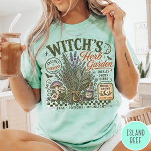 May include: A light mint green t-shirt featuring a graphic print with the text Witch's Herb Garden, Freshly Gathered, Locally Grown Herbs, Moonlit Grown, Autumn Picked, Sage, Potions, Moonlight, and Witch's Brew, alongside illustrations of herbs, mushrooms, and a bottle.