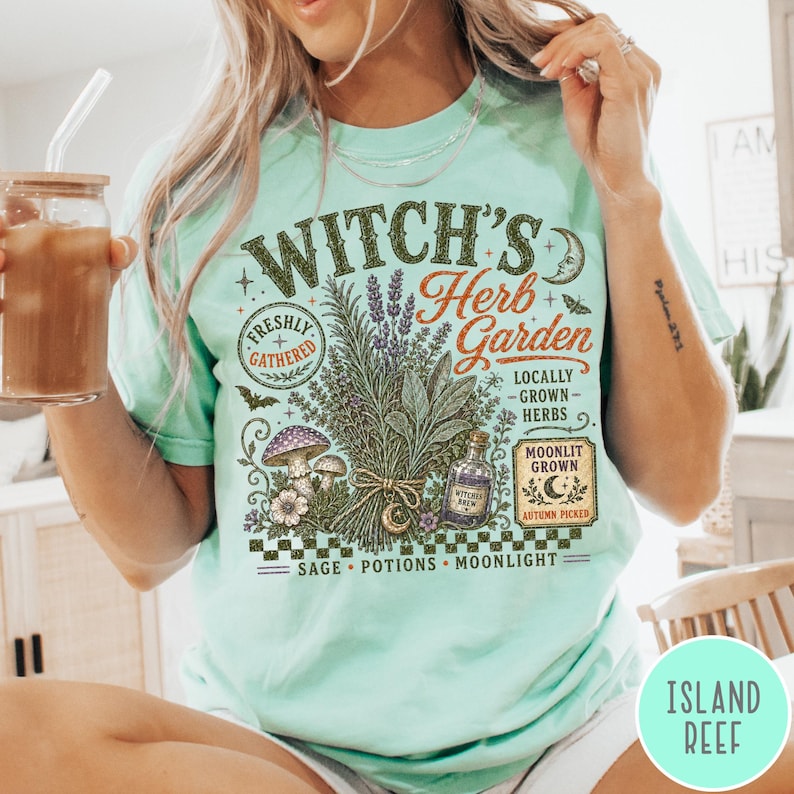 May include: A light mint green t-shirt featuring a graphic print with the text Witch's Herb Garden, Freshly Gathered, Locally Grown Herbs, Moonlit Grown, Autumn Picked, Sage, Potions, Moonlight, and Witch's Brew, alongside illustrations of herbs, mushrooms, and a bottle.