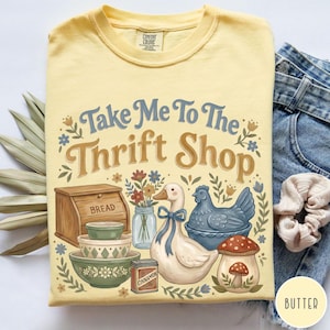 Op de afbeelding: Geel Comfort Colors T-shirt met een afbeelding met de tekst "Take Me To The Thrift Shop". De afbeelding bevat een brooddoos, kommen, een eend, een kip en een paddenstoel. Het T-shirt is opgevouwen.