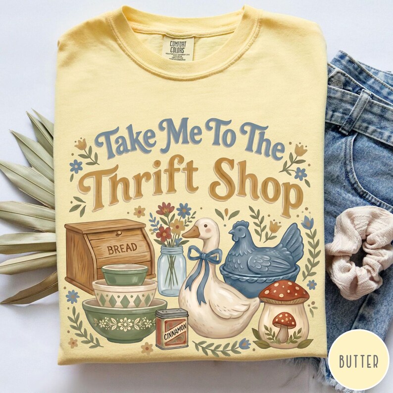 Op de afbeelding: Geel Comfort Colors T-shirt met een afbeelding met de tekst "Take Me To The Thrift Shop". De afbeelding bevat een brooddoos, kommen, een eend, een kip en een paddenstoel. Het T-shirt is opgevouwen.
