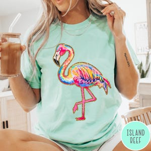 May include: A light green t-shirt featuring a colorful flamingo design. The flamingo is depicted with vibrant rainbow-colored feathers, a pink body, and a gold glitter outline. The shirt has a relaxed fit.