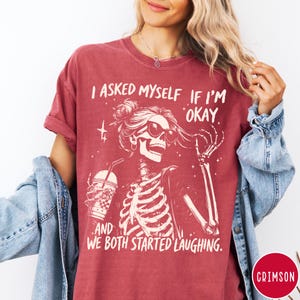 May include: Burgundy t-shirt with a skeleton graphic wearing sunglasses and holding a drink. The text on the shirt reads, "I asked myself if I'm okay, and we both started laughing."
