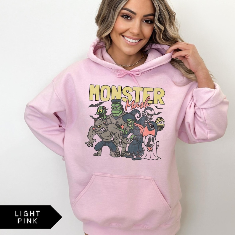 Puede incluir: Una sudadera con capucha rosa claro con un estampado gr&aacute;fico de un grupo de monstruos. El texto "Monster Mash" est&aacute; impreso en amarillo sobre el gr&aacute;fico.