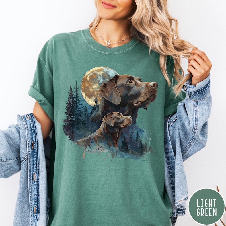 May include: Light green t-shirt featuring a watercolor illustration of two brown dogs with a full moon and forest background. The shirt has a relaxed fit and a crew neck. The words "LIGHT GREEN" are printed on a green circle.