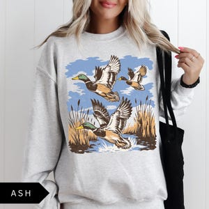 May include: Ash gray sweatshirt featuring a graphic of three ducks in flight over water and reeds. The ducks have brown and white wings, green heads, and yellow beaks. The background is blue with stylized clouds.