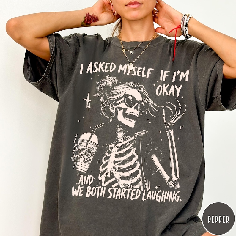 May include: Dark gray t-shirt with a skeleton graphic wearing sunglasses and holding a drink. The text on the shirt reads, "I asked myself if I'm okay, and we both started laughing."