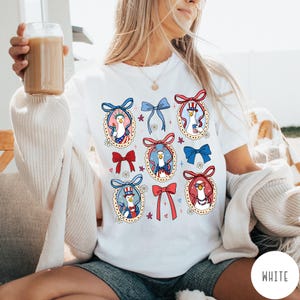 May include: White t-shirt featuring a patriotic design with cartoon geese in oval frames, adorned with red, white, and blue bows. The geese are dressed in festive attire, including a top hat and sunglasses. The shirt has the word "WHITE" printed on the bottom right.
