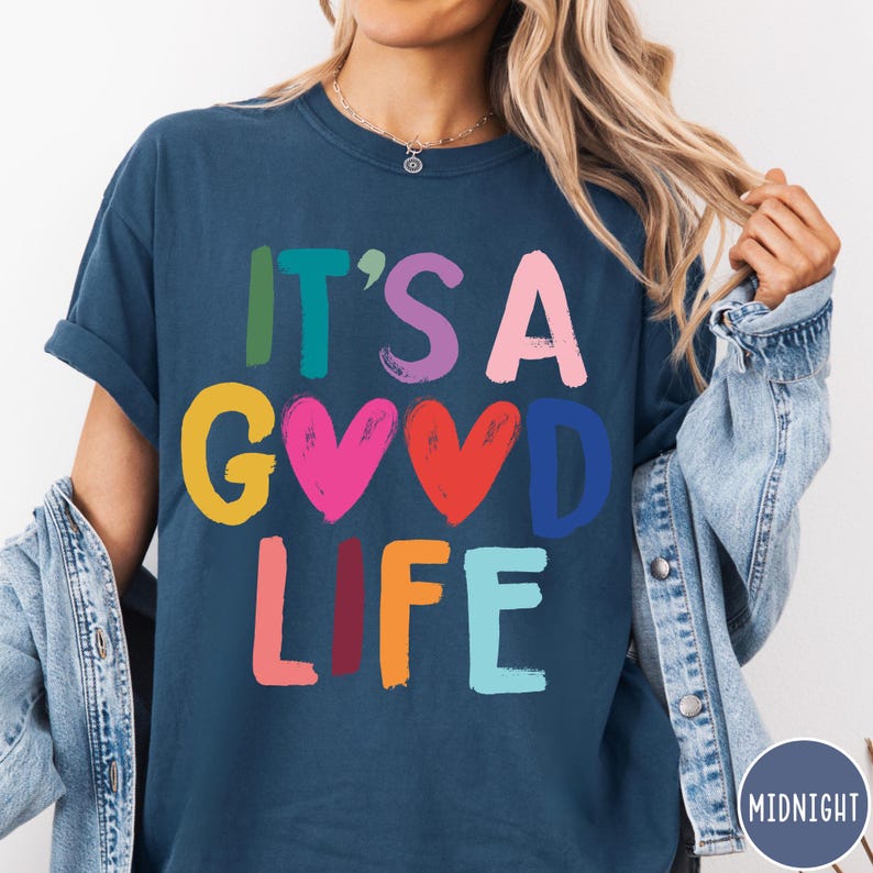 K&ouml;nnte beinhalten: Ein dunkelblaues T-Shirt mit kurzen &Auml;rmeln und dem mehrfarbigen Schriftzug "IT'S A GOOD LIFE", bei dem zwei rote und rosa Herzen den Buchstaben O ersetzen. Die Grafik verwendet einen Pinselstrich-Schriftstil mit Buchstaben in Gr&uuml;n, Blau, Lila, Pink, Gelb und Orange.