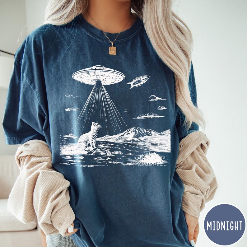Pode incluir: Uma camiseta azul marinho com um gr&aacute;fico branco de um gato olhando para cima para um OVNI. O OVNI est&aacute; emitindo raios de luz e uma pequena nave espacial est&aacute; voando acima. O gr&aacute;fico est&aacute; sobre um fundo branco com um padr&atilde;o ondulado na parte inferior.