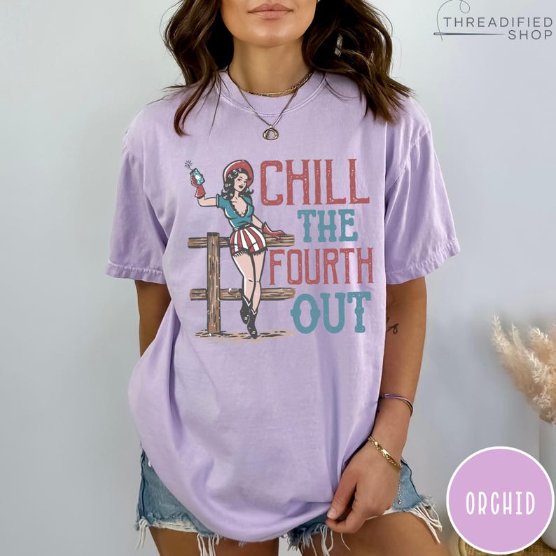 May include: A light purple t-shirt with the text "CHILL THE FOURTH OUT" and a graphic of a woman in patriotic attire. The woman is holding a sparkler. The shirt is a comfortable, casual style.