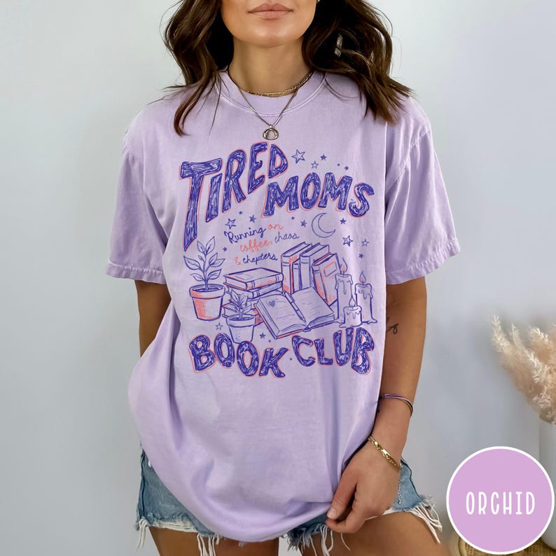 May include: A light purple t-shirt with the text "Tired Moms Book Club" in a whimsical font. The graphic includes illustrations of books, plants, and candles. The shirt also has the phrase "Running on coffee, chaos & chapters".